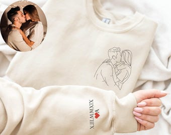 May include: A beige crewneck sweatshirt featuring a black line art illustration of a couple kissing on the chest. The sleeve is embroidered with the Roman numeral date X.III.MMXIX, a red heart icon, and the letter A in black thread.