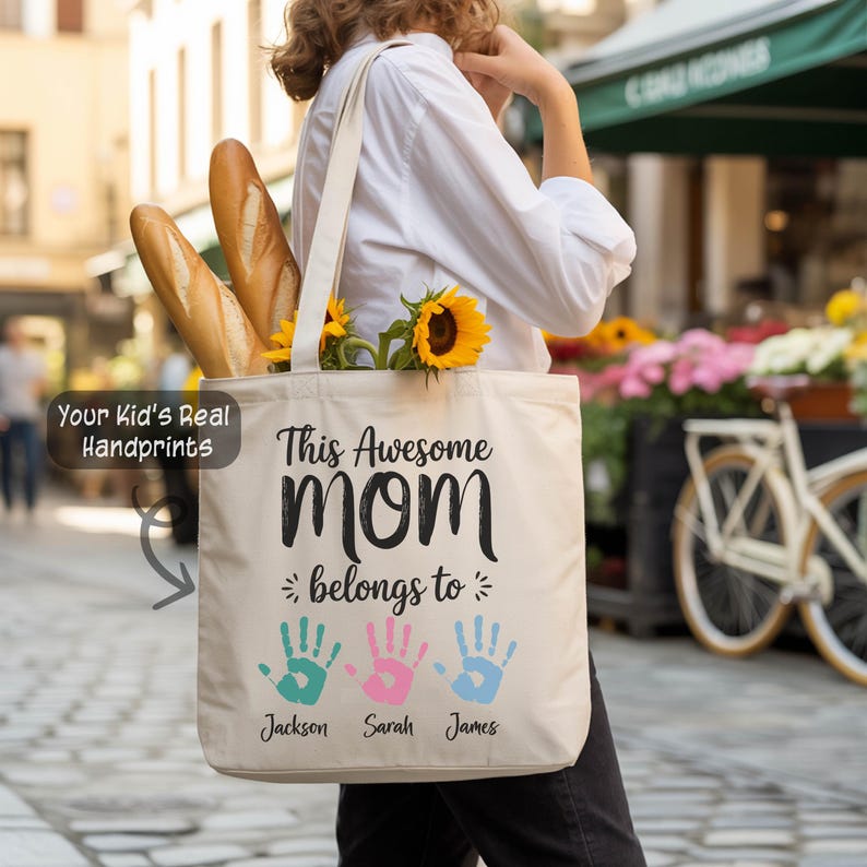 A person is walking down a street carrying a white tote bag with a printed message that says "This Awesome Mom belongs to Jackson, Sarah, and James".