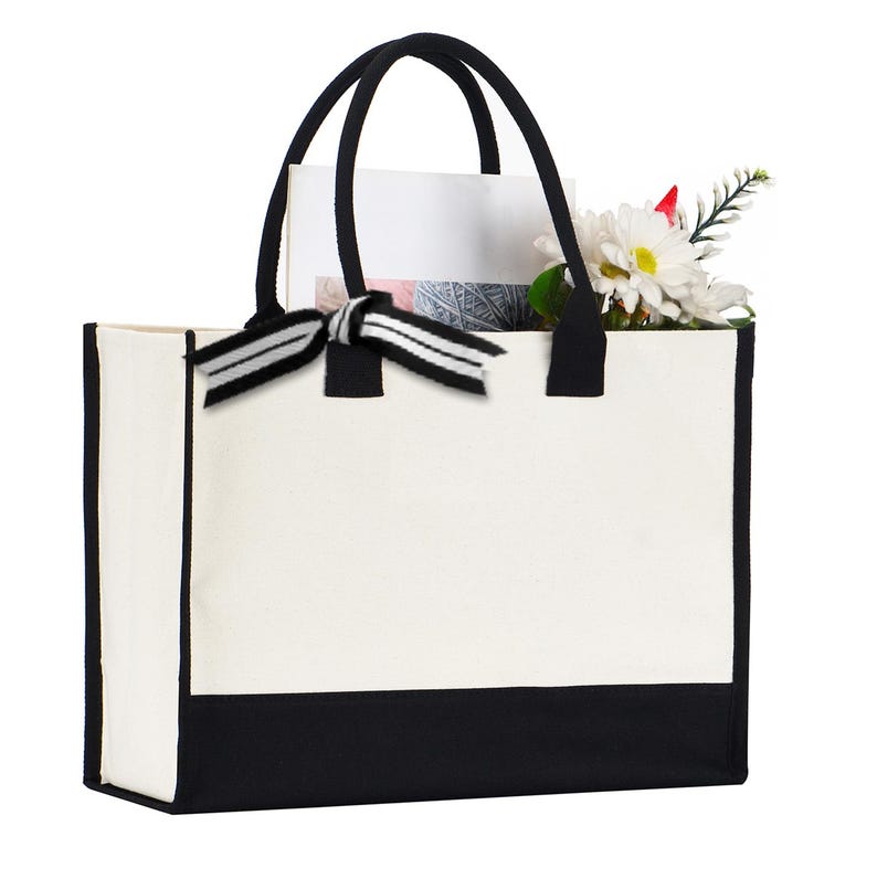 A white tote bag with a black trim, featuring a black bow tied at the top. The bag is holding a bouquet of flowers, including a white daisy and a red flower.