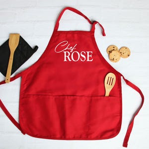 May include: A red apron with the text "Chef ROSE" in white script. A wooden spoon and a black oven mitt are on the left. Three chocolate chip cookies are on the right. The apron has two pockets.