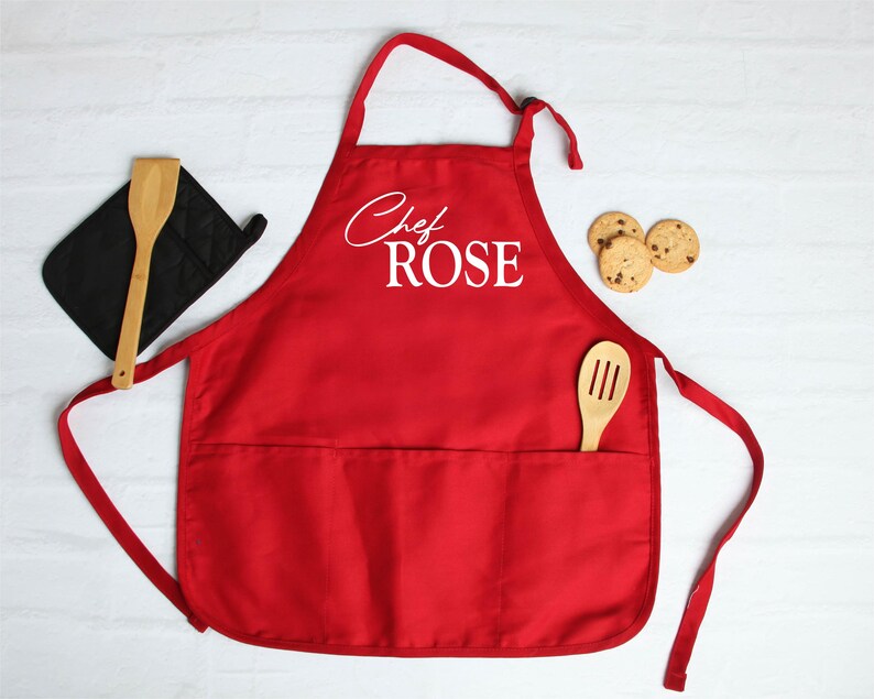 May include: A red apron with the text "Chef ROSE" in white script. A wooden spoon and a black oven mitt are on the left. Three chocolate chip cookies are on the right. The apron has two pockets.