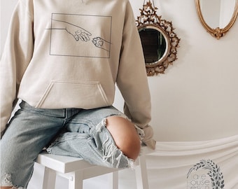 May include: A beige hooded sweatshirt featuring a black line art graphic of a human hand reaching toward a dog paw inside a rectangular frame. The hoodie includes long sleeves, a front kangaroo pocket, and a drawstring hood with matching beige cords.