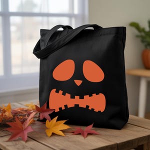 A black tote bag with an orange pumpkin face design sits on a wooden table, surrounded by fallen autumn leaves.