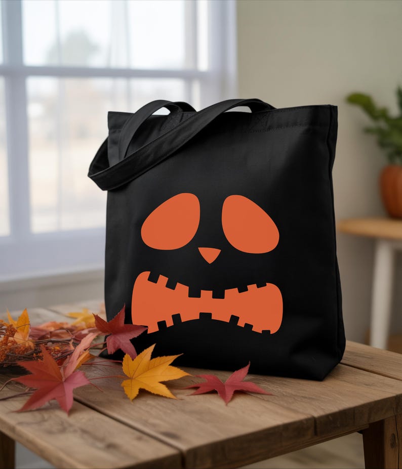 A black tote bag with an orange pumpkin face design sits on a wooden table, surrounded by fallen autumn leaves.