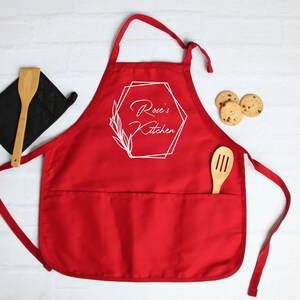 May include: Red kitchen apron with a large pocket and adjustable straps. The apron has a white geometric design with the text "Rose's Kitchen" in a script font.