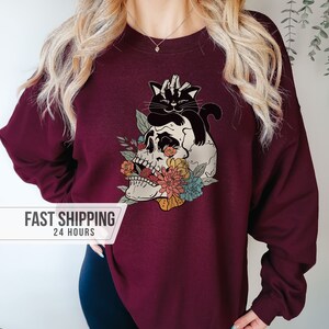 May include: Burgundy sweatshirt featuring a graphic of a skull with floral accents and a black cat. The skull has colorful flowers and the cat is perched on top. The sweatshirt is a solid color.