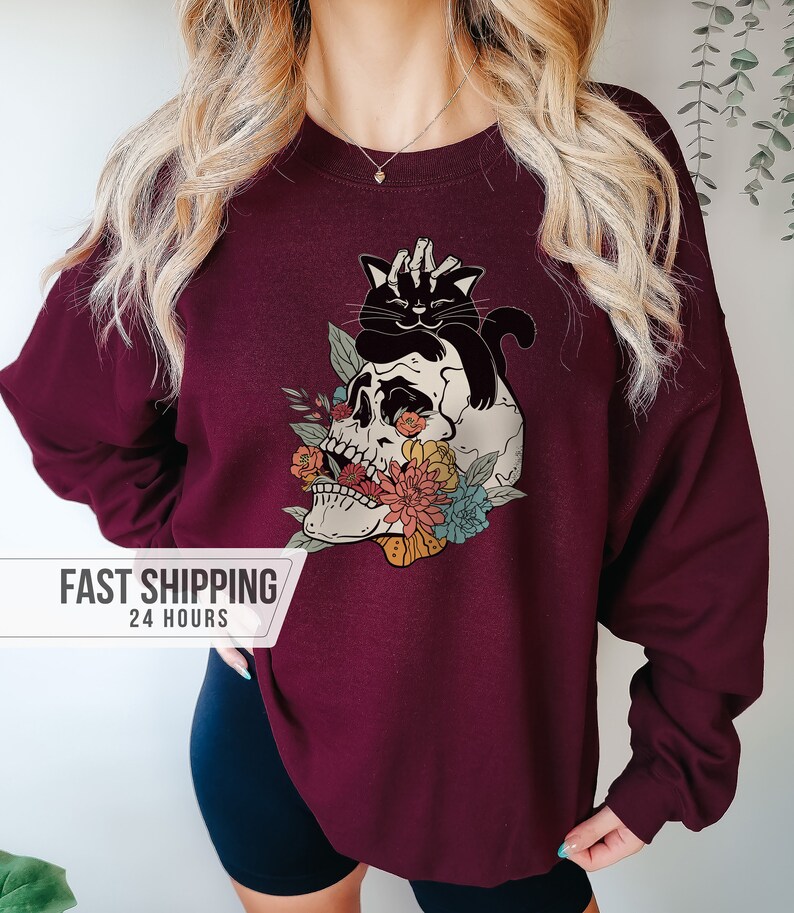 May include: Burgundy sweatshirt featuring a graphic of a skull with floral accents and a black cat. The skull has colorful flowers and the cat is perched on top. The sweatshirt is a solid color.