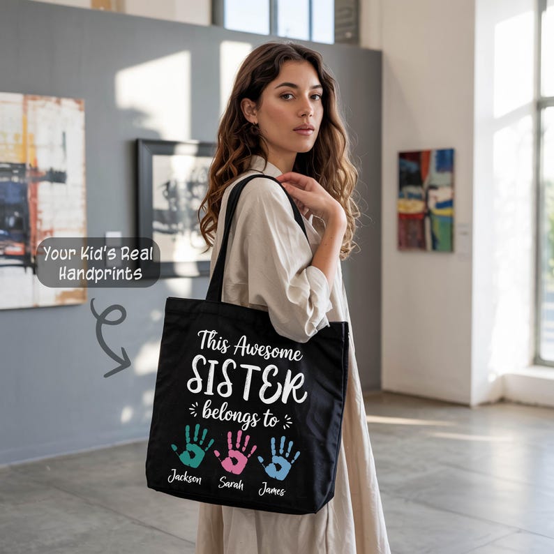 A woman wearing a beige dress stands in an art gallery, holding a black tote bag with a personalized message printed on it.
