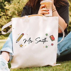 May include: Cream-colored tote bag with the name "Mr. Smith" in black script. The bag features a yellow pencil, a red book, and colorful star accents. The bag has white handles and is made of a sturdy fabric.
