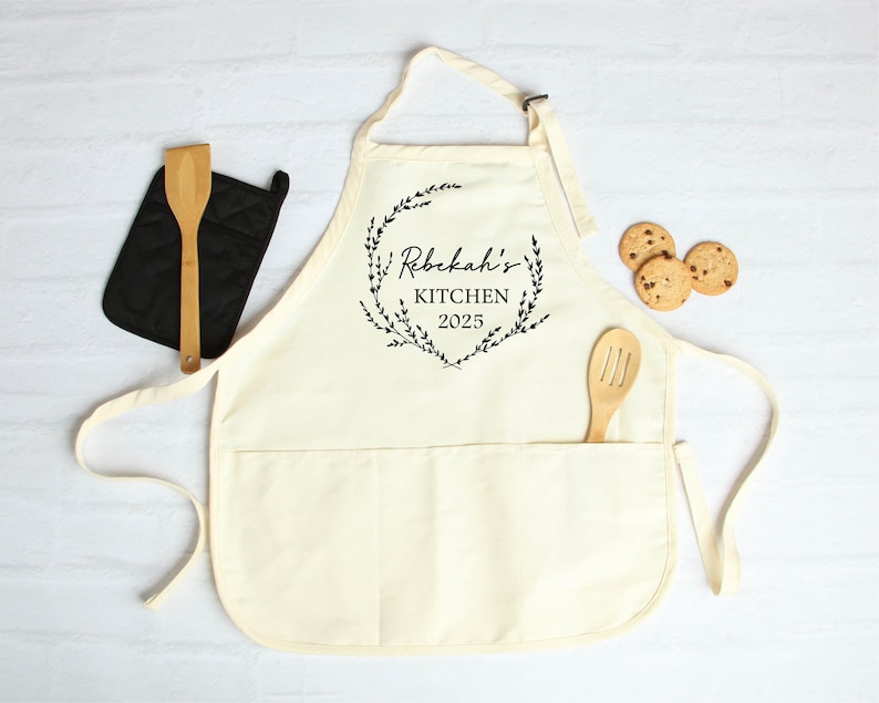 K&ouml;nnte beinhalten: Eine wei&szlig;e Baumwollsch&uuml;rze mit einer gro&szlig;en Tasche und verstellbaren Tr&auml;gern. Die Sch&uuml;rze hat einen schwarzen Aufdruck einer Blumengirlande mit dem Text "Rebekah's Kitchen 2025" darin.