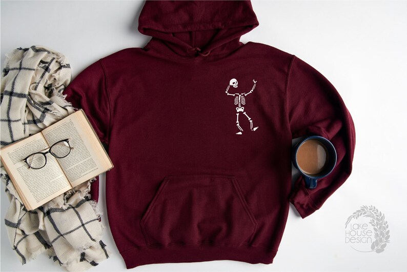 K&ouml;nnte beinhalten: Ein weinroter Kapuzenpullover mit einem wei&szlig;en Skelett-Motiv auf der Vorderseite. Der Hoodie hat eine K&auml;ngurutasche und eine Kapuze mit Kordelzug. Ein Buch, eine Brille und ein Schal liegen neben dem Hoodie.