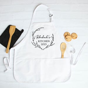 K&ouml;nnte beinhalten: Wei&szlig;e K&uuml;chensch&uuml;rze mit einem schwarzen floralen Kranzdesign und dem Text "Rebekah's Kitchen 2023".