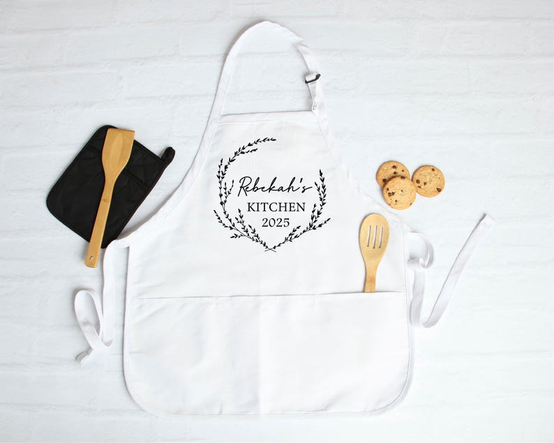 K&ouml;nnte beinhalten: Wei&szlig;e K&uuml;chensch&uuml;rze mit einem schwarzen floralen Kranzdesign und dem Text "Rebekah's Kitchen 2023".