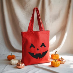 A red tote bag with a black jack-o-lantern face design sits on a white surface, surrounded by small pumpkins.