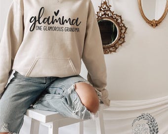 Könnte beinhalten: Ein beigefarbener Kapuzenpullover mit dem schwarzen Schriftzug glamma und dem Satz ONE GLAMOROUS GRANDMA sowie einem kleinen Herzsymbol über dem Text. Der Hoodie verfügt über eine Kängurutasche vorne, lange Ärmel und eine Kapuze mit Kordelzug.