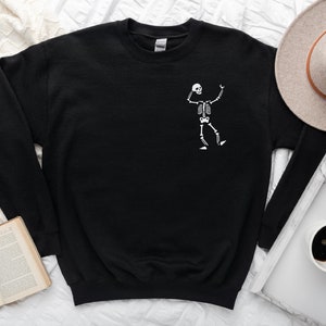 K&ouml;nnte beinhalten: Schwarzes Sweatshirt mit einem wei&szlig;en Skelett-Design auf der Vorderseite. Das lang&auml;rmlige Oberteil mit Rundhalsausschnitt ist aus weichem Material. Ein Hut, eine Kaffeetasse und ein Buch befinden sich im Hintergrund.