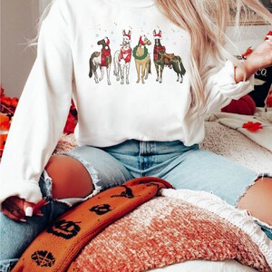 May include: White long-sleeve sweatshirt featuring a festive design of five horses adorned with Christmas decorations. Each horse is depicted with a unique combination of hats, scarves, and saddle blankets, creating a holiday theme. The design includes snowflakes and gold accents.
