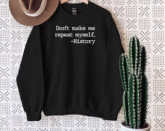 Pode incluir: Uma camisola preta de manga comprida e gola redonda com o texto impresso a branco "Don't make me repeat myself. -History" centrado no peito. A peça é exibida num cabide de madeira, acompanhada por um chapéu fedora castanho.
