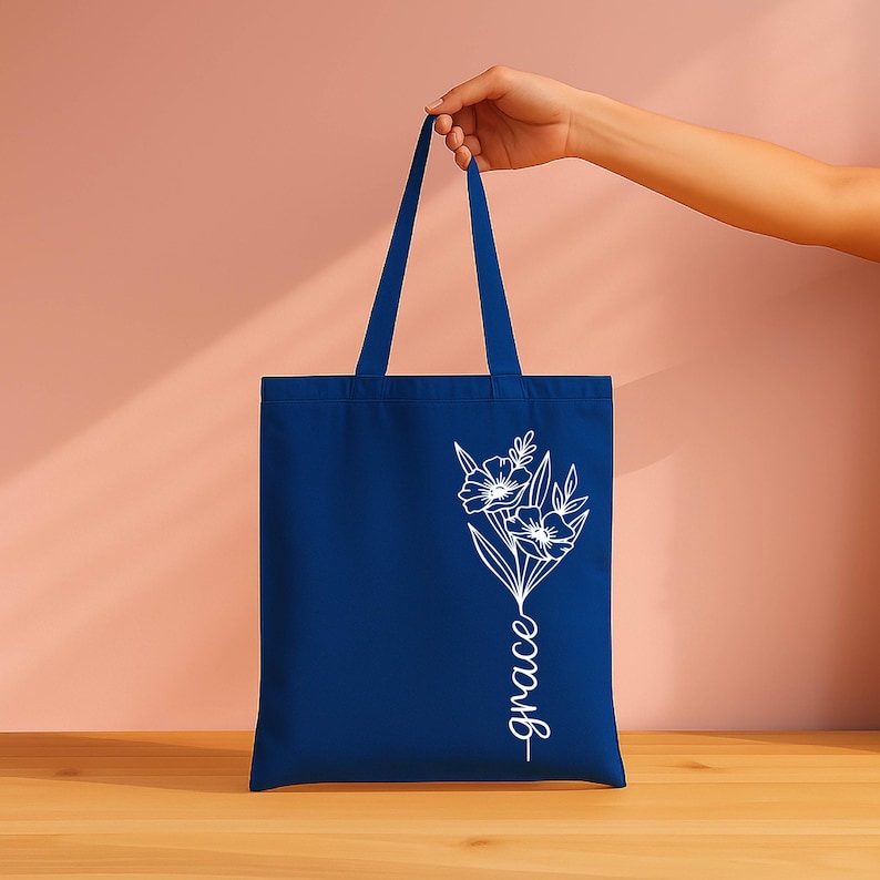 A hand holding a blue tote bag with a white floral design on it, against a pink background.