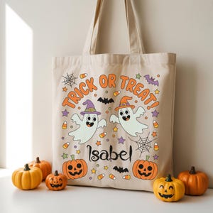 May include: Beige canvas tote bag with a Halloween-themed design. The bag features the words "Trick or Treat!" in orange, along with two cartoon ghosts wearing witch hats, pumpkins, bats, and the name "Isabel".