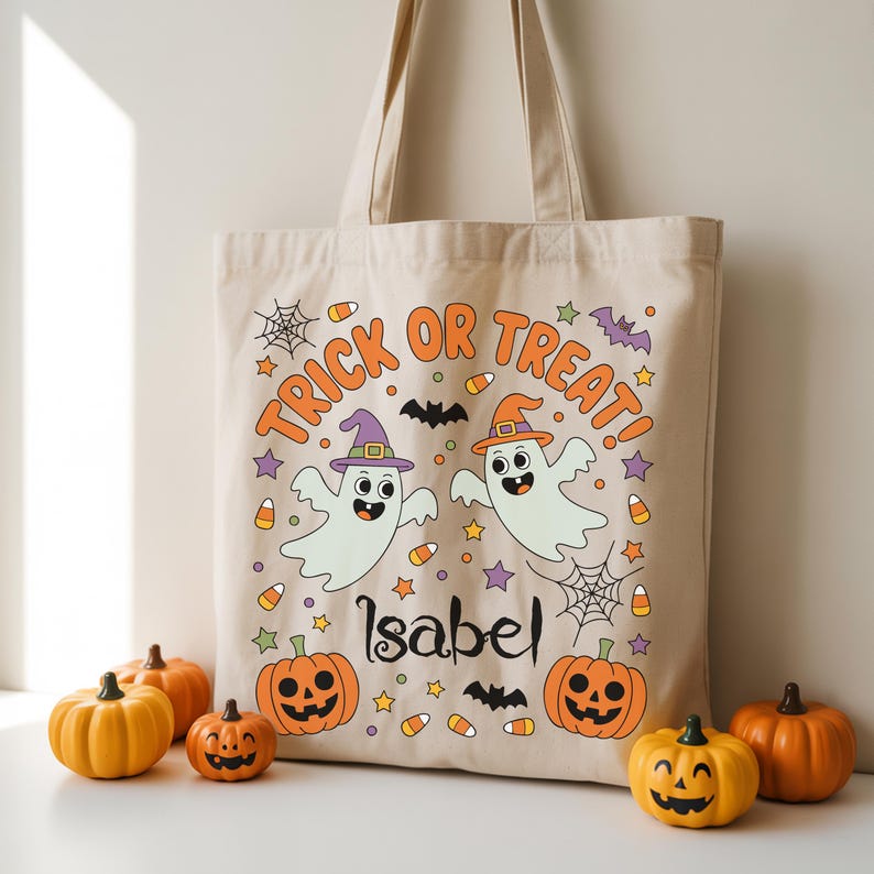May include: Beige canvas tote bag with a Halloween-themed design. The bag features the words "Trick or Treat!" in orange, along with two cartoon ghosts wearing witch hats, pumpkins, bats, and the name "Isabel".