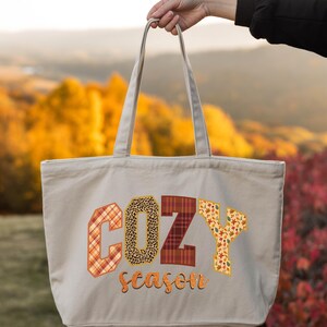 A hand holding a large tote bag with the word "COZY" printed on it, against a backdrop of autumn foliage.