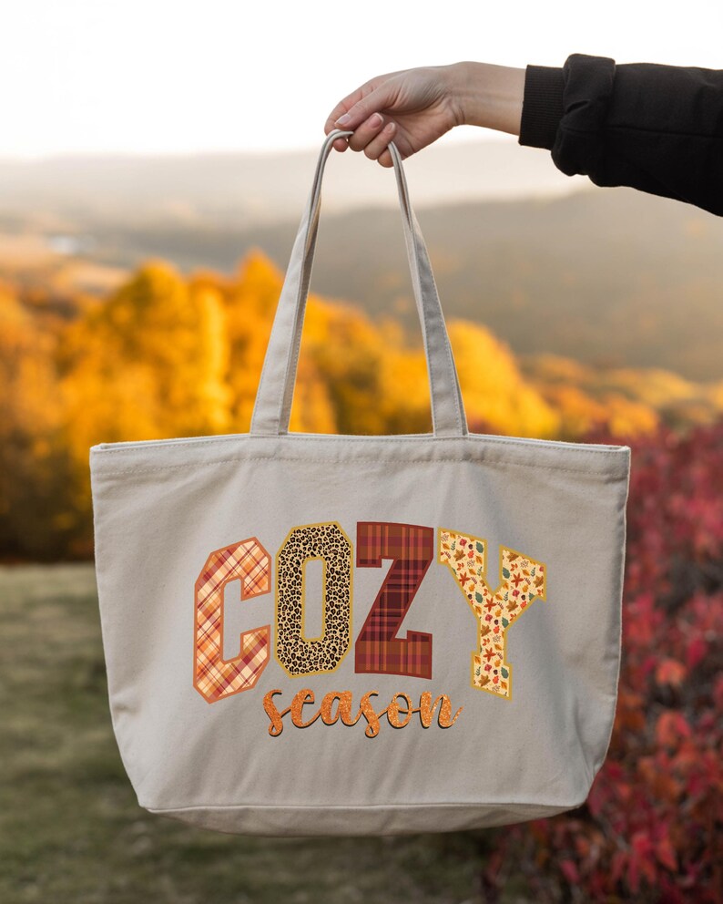A hand holding a large tote bag with the word "COZY" printed on it, against a backdrop of autumn foliage.