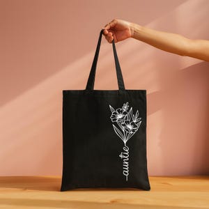 A hand holding a black tote bag with a white floral design on it, against a pink background.