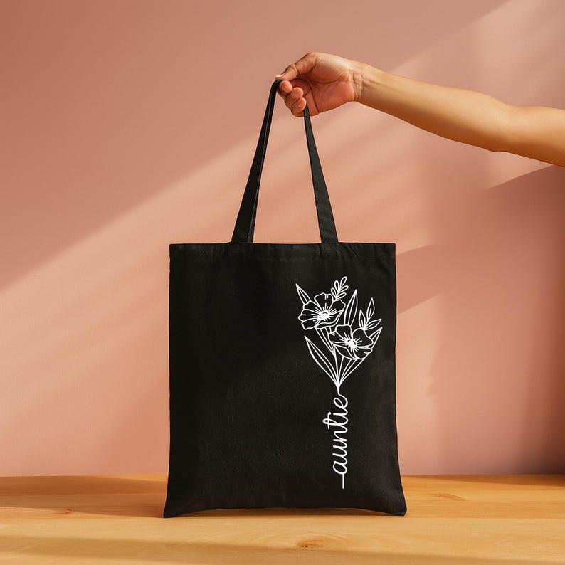 A hand holding a black tote bag with a white floral design on it, against a pink background.