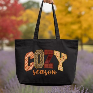 A hand holding a black tote bag with the word "COZY" printed on it, against a background of colorful autumn foliage.