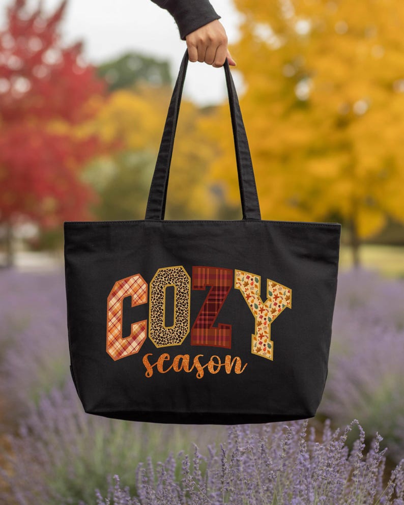 A hand holding a black tote bag with the word "COZY" printed on it, against a background of colorful autumn foliage.