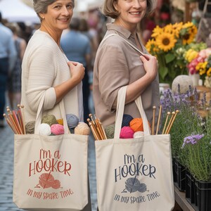May include: Two women holding beige tote bags filled with yarn, knitting needles, and colorful balls of yarn. The bags have the text "I'm a Hooker in my spare time" printed on them in red and navy blue.