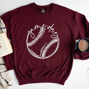 May include: A maroon sweatshirt with a white baseball graphic and the text "game day" in a handwritten font.