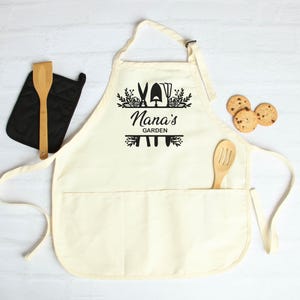May include: A white cotton apron with a large pocket and adjustable neck straps. The apron has a black design of gardening tools and the text "Nana's Garden".