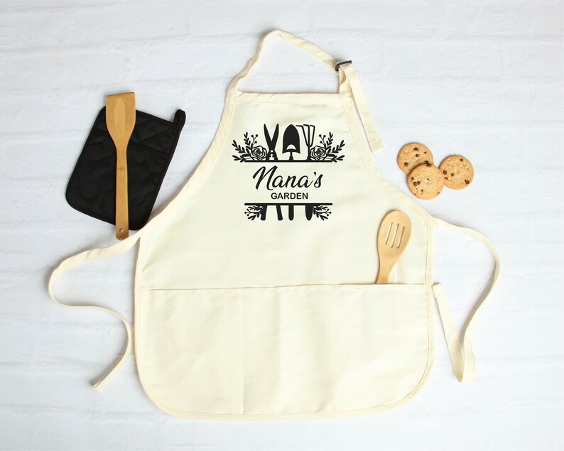 May include: A white cotton apron with a large pocket and adjustable neck straps. The apron has a black design of gardening tools and the text "Nana's Garden".
