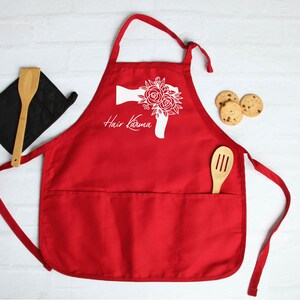 May include: Red kitchen apron with a large white graphic of a hairdryer with flowers. The text "Hair Karma" is printed below the graphic. The apron has a large pocket in the front.