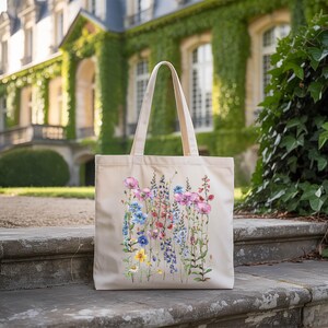 A beige tote bag with a floral design is placed on a stone step in front of a building with ivy growing on it.