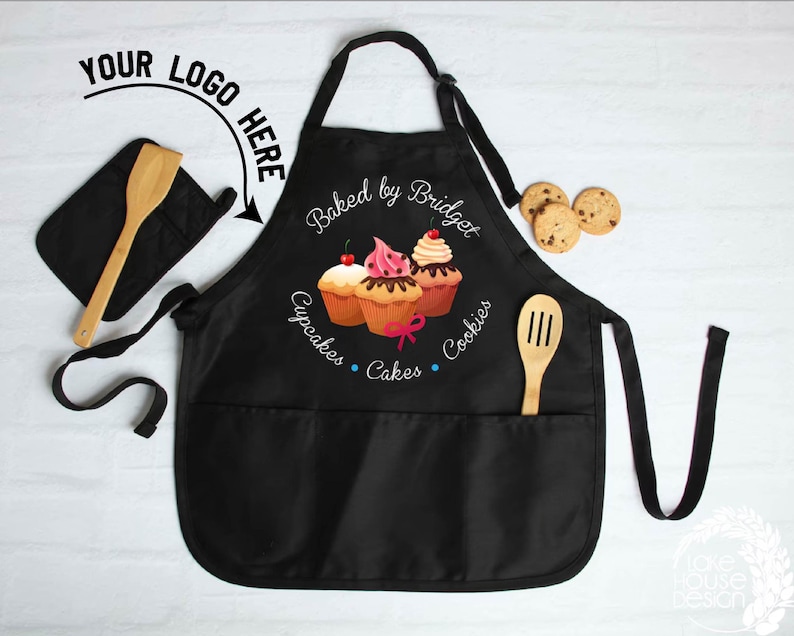 May include: Black apron with two pockets, a wooden spoon, and a matching oven mitt. The apron features a cupcake design with the text "Baked by Bridget" and the words "Cupcakes, Cakes, Cookies". A few chocolate chip cookies are also visible.