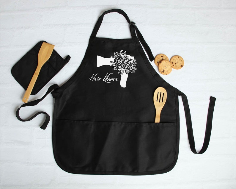 May include: Black kitchen apron with a white floral hairdryer design and the text "Hair Karma". The apron has two pockets and adjustable straps.