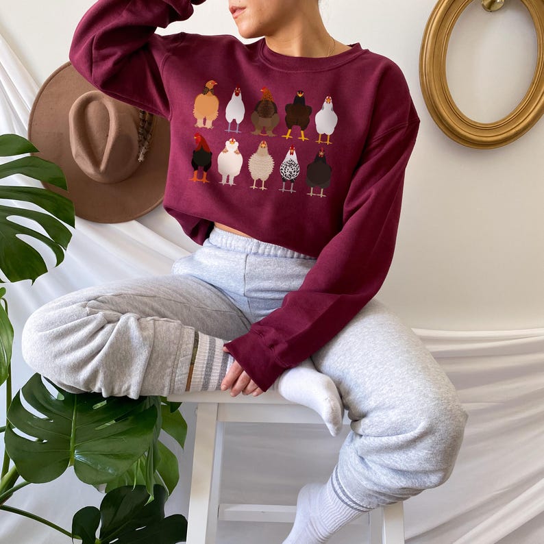 May include: A maroon cropped sweatshirt with a graphic print of a group of chickens. The sweatshirt is worn by a person wearing grey sweatpants and white socks.