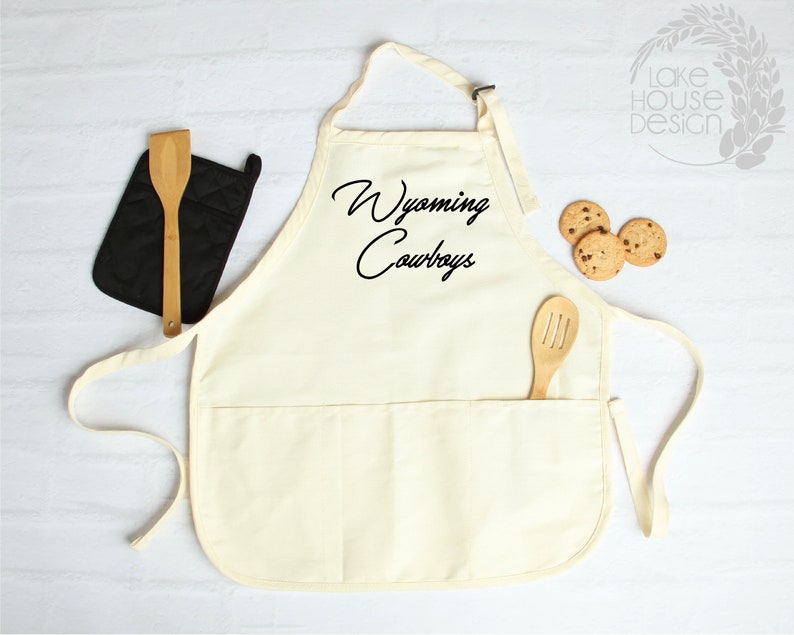 May include: A white cotton apron with a large pocket and adjustable straps. The apron has the text "Wyoming Cowboys" printed in black on the front.