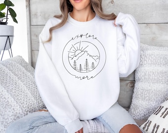 Könnte beinhalten: Ein weißes Sweatshirt mit Rundhalsausschnitt und einer schwarzen Strichgrafik auf der Brust. Das Design besteht aus einem kreisförmigen Rahmen, der ein Gebirge, Tannen und eine aufgehende Sonne umschließt, mit dem Text "explore more" in Schreibschrift.