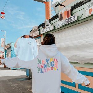 May include: White hoodie with a colorful graphic that says "Never Enough Plants" with a flower design. The hoodie is worn by a person standing in front of a building with a sign that says "Ice Cold Beer".