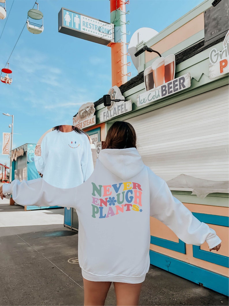 May include: White hoodie with a colorful graphic that says "Never Enough Plants" with a flower design. The hoodie is worn by a person standing in front of a building with a sign that says "Ice Cold Beer".