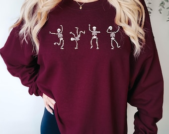 May include: A long-sleeve crewneck sweatshirt in a deep maroon color featuring a white graphic print of four distinct skeletons in various dancing poses arranged in a horizontal line across the center of the chest area of the garment.