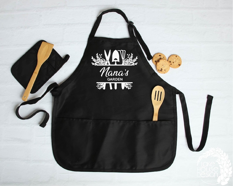 May include: Black gardening apron with two pockets and adjustable straps. The apron has a white design with gardening tools and the text "Nana's Garden".