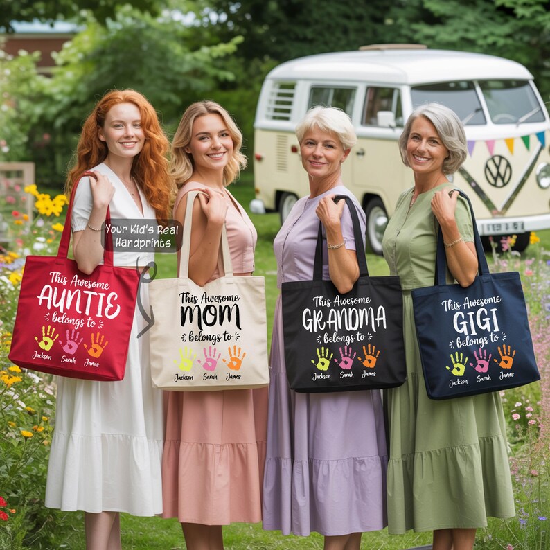 Four women standing in a row, each holding a tote bag with a handprint design. The tote bags are in different colors and have text that says "This Awesome Auntie Belongs to" and "This Awesome Mom Belongs to". In the background, there is a Volkswagen van.