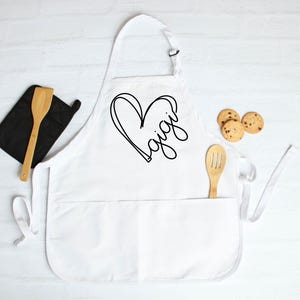 May include: White apron with a black heart and the word "Gigi" written inside. The apron has two pockets and adjustable straps.