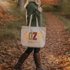A woman wearing a green jacket and boots is walking on a path through a forest with fallen leaves. She is carrying a large tote bag with the word "COZY season" printed on it.