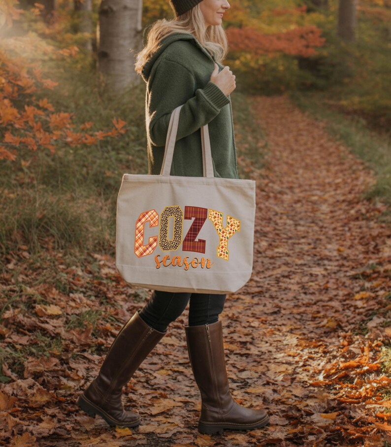 A woman wearing a green jacket and boots is walking on a path through a forest with fallen leaves. She is carrying a large tote bag with the word "COZY season" printed on it.